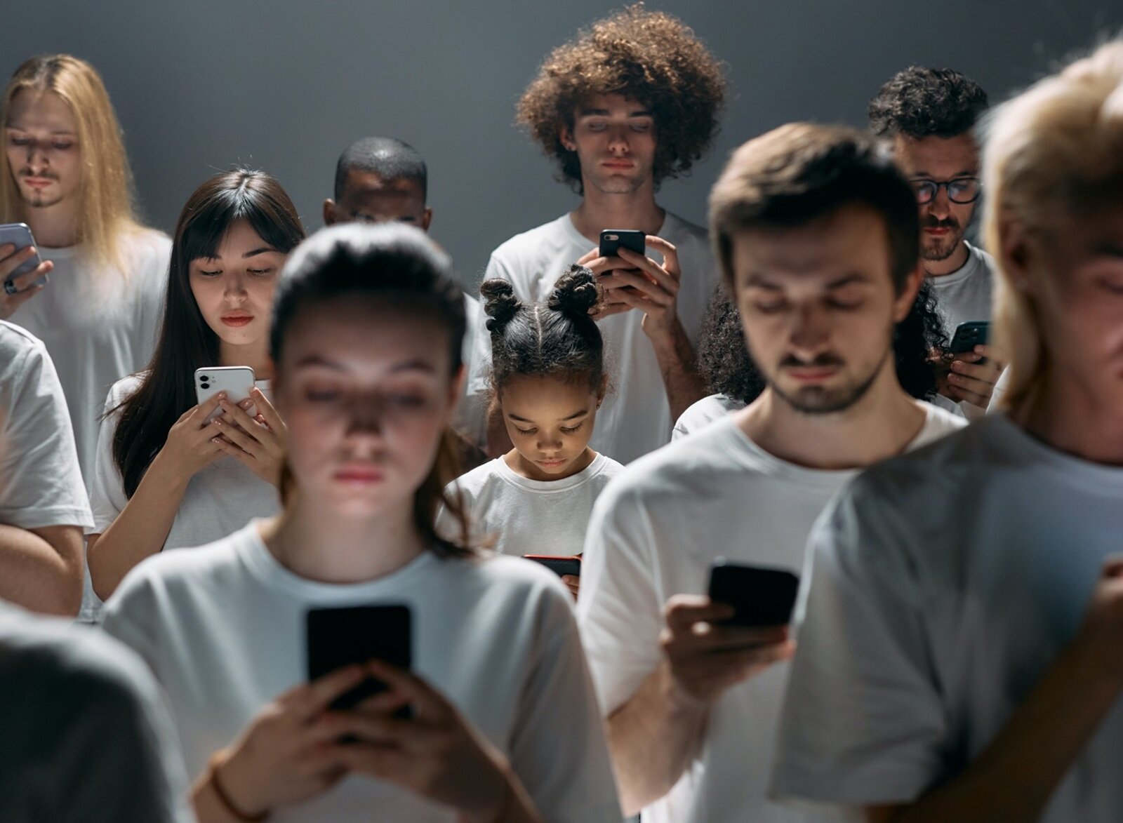 Zdjęcie ukazuje grupę młodych osób, w ujęciu od pasa w górę, które stoją w kilku rzędach. Każda osoba trzyma telefon smartfon, ma lekko pochyloną głowę i spogląda na ekran telefonu. Wszystkie osoby ubrane są w białe koszulki z krótkim rękawem.