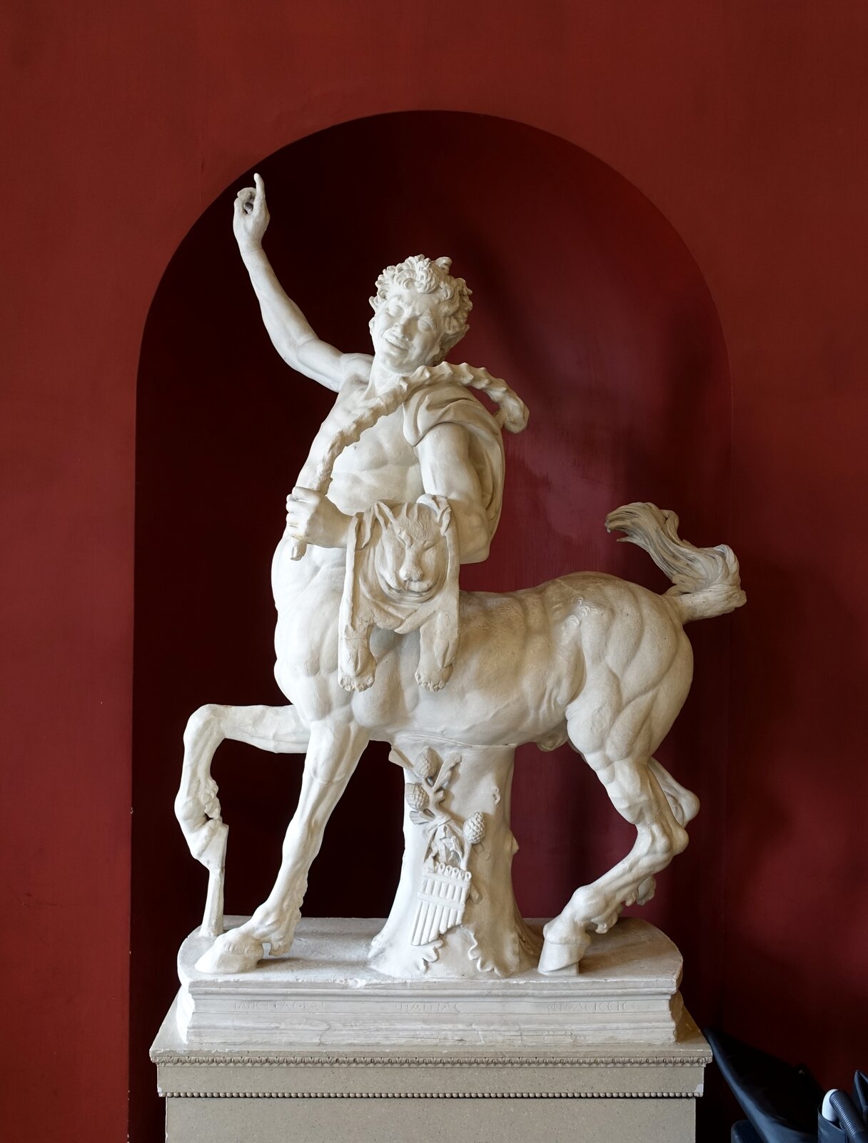 Na zdjęciu zaprezentowano starożytną rzeźbę, która mieści się w pomieszczeniu o czerwonych ścianach. Statua jest koloru białego i przedstawia centaura. Głowa wraz z klatką piersiową oraz rękoma należą do człowieka, natomiast pozostała jego część należy do konia. Postać ma uniesioną prawą rękę, z wysuniętym wskazującym palcem, skierowanym w stronę nieba, a twarz obrócona jest w lewą stronę. Centaur w swej lewej dłoni trzyma bicz, a przez ramie przewieszoną ma skórę ze zwierzęcia.