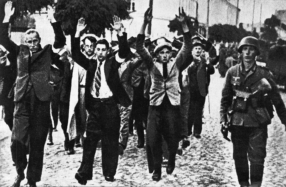 Zdjęcie przedstawia grupę ludzi. Brukowaną ulicą idzie grupa mężczyzn. Mają podniesiono wysoko ręce. Dwóch mężczyzn na przedzie ma białe przepaski na ramionach. Mężczyzn eskortuje niemiecki żołnierz. Jest w mundurze i hełmie. Na prawym ramieniu ma zawieszoną broń. 