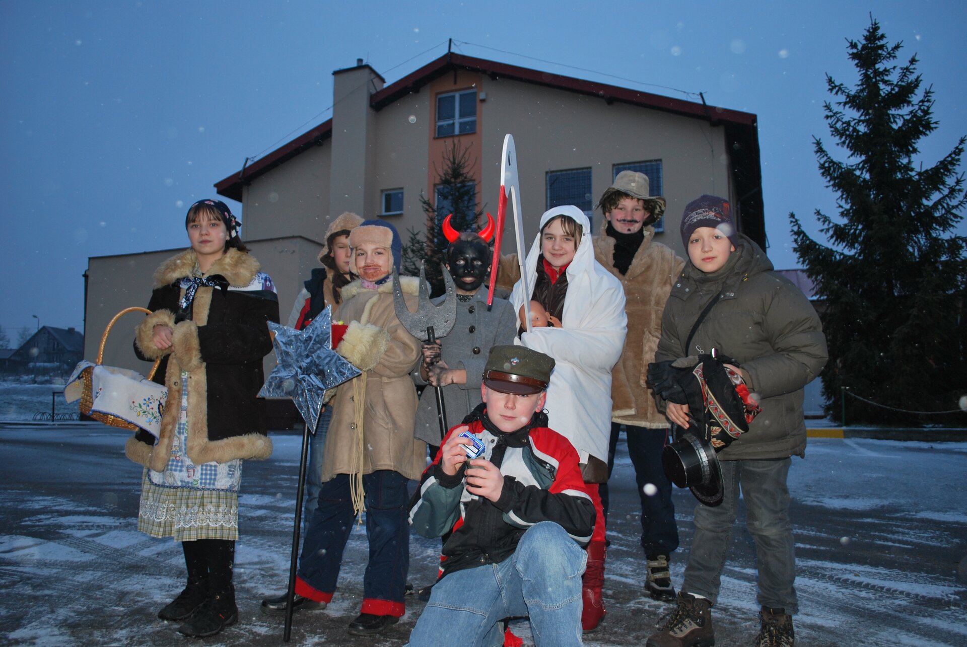 Kliknij, aby powiększyć. Zdjęcie przedstawia kolędników. Ośmioro dzieci przebranych jest w różne stroje: jeden z chłopców nosi czarną maskę z rogami i trzyma widły, drugi chłopiec ma wojskową czapkę, dziewczynka przebrana jest w kożuch i spódnicę, trzyma koszyk. Chłopiec z narzuconym na siebie białym prześcieradłem trzyma głowę bociana zrobioną z drewna i pomalowaną. Jedno z dzieci trzyma gwiazdę. W tle budynek, drzewa i ciemniejące niebo.