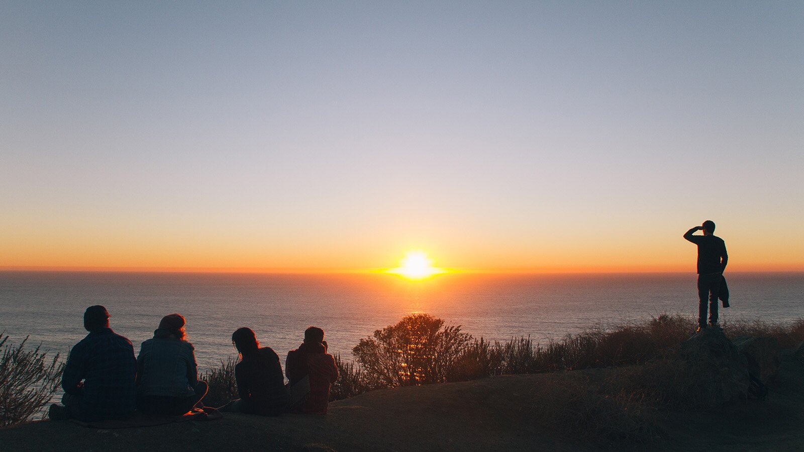 Zdjęcie przedstawia cztery osoby siedzące na wydmach oraz piątą stojącą nieopodal. Wszyscy patrzą w stronę morza, a na horyzoncie jest słońce zlewające się z jego taflą.