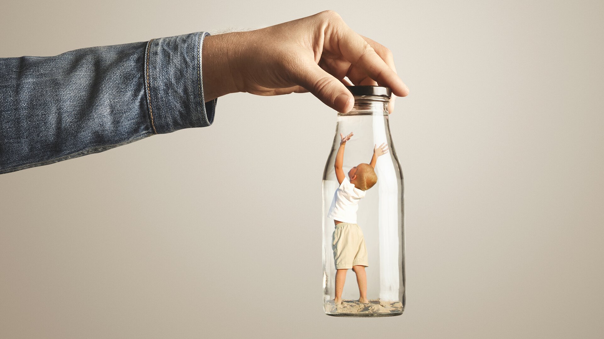 Zdjęcie przedstawia przykładowy fotomontaż. Po lewej stronie widoczna jest ręka od przedramienia, w jeansowej koszuli. Ręka trzyma przeźroczystą butelkę z metalową nakrętką, w środku której widoczne jest małe dziecko, zwrócone w stronę ręki trzymającej butelkę. Dziecko ma obydwie ręce podniesione do góry, jest ubrane w białą koszulkę i beżowe szorty, i stoi na piasku.