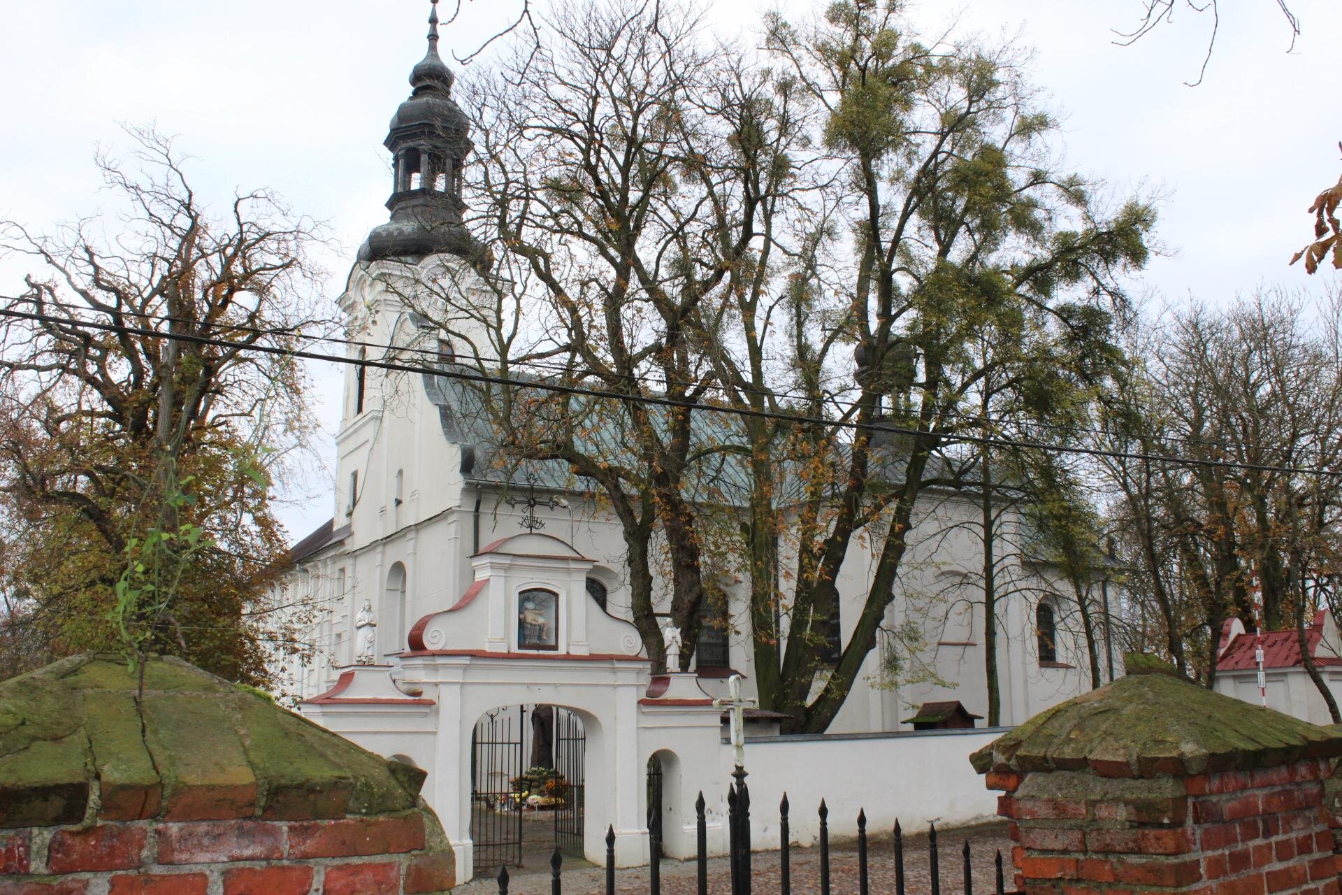 Zdjęcie przedstawia biały budynek widziany z boku. Na pierwszym planie znajduje się fragment ceglano‑metalowego płotu. Kilka metrów dalej widać ozdobną białą bramę i betonowe lub murowane ogrodzenie w tym samym kolorze. Za bramą znajduje się budynek z wieżą, na szczycie której widoczny jest szpikulec. Bryłę budynku częściowo przysłaniają drzewa
