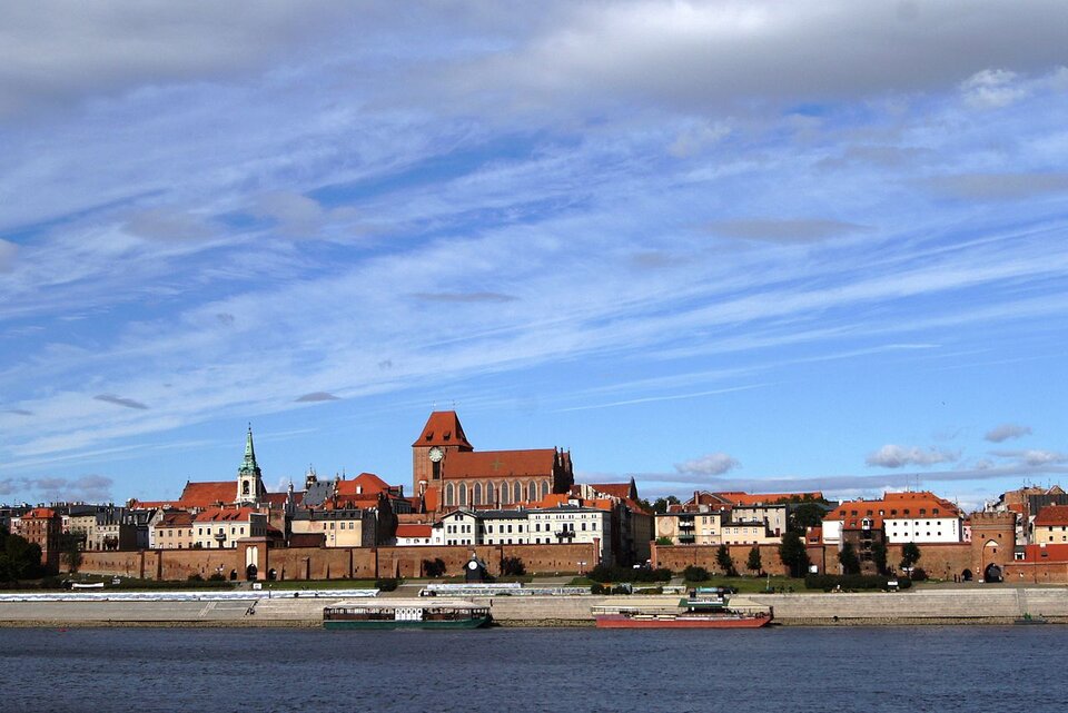 Zdjęcie przedstawia panoramę miasta z drugiego brzegu rzeki. To średniowieczny zespół miejski Torunia. Budynki stoją za murami miasta. Mają czerwone dachy. Większość z nich wykonana jest z czerwonej cegły. Wyróżnia się wysoki budynek gotyckiej katedry. Na pierwszym planie fotografii jest rzeka. 