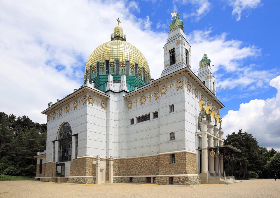 Kościół św. Leopolda na Steinhof w Wiedniu, zaprojektowany przez Otto Wagnera. Monumentalny biały budynek ma dużą, pokrytą złotymi płytkami kopułę zwieńczoną krzyżem oraz dwie smukłe wieże z dekoracyjnymi figurami. Elewację zdobią złote ornamenty, krzyże i geometryczne detale. Po prawej stronie znajduje się portyk wsparty na wysokich kolumnach, ozdobiony złotymi elementami. Dolna część ścian jest wyłożona nieregularnym kamieniem. Kościół otaczają drzewa, a przed nim znajduje się szeroki, jasny plac. Nad budynkiem widać błękitne niebo z białymi chmurami.