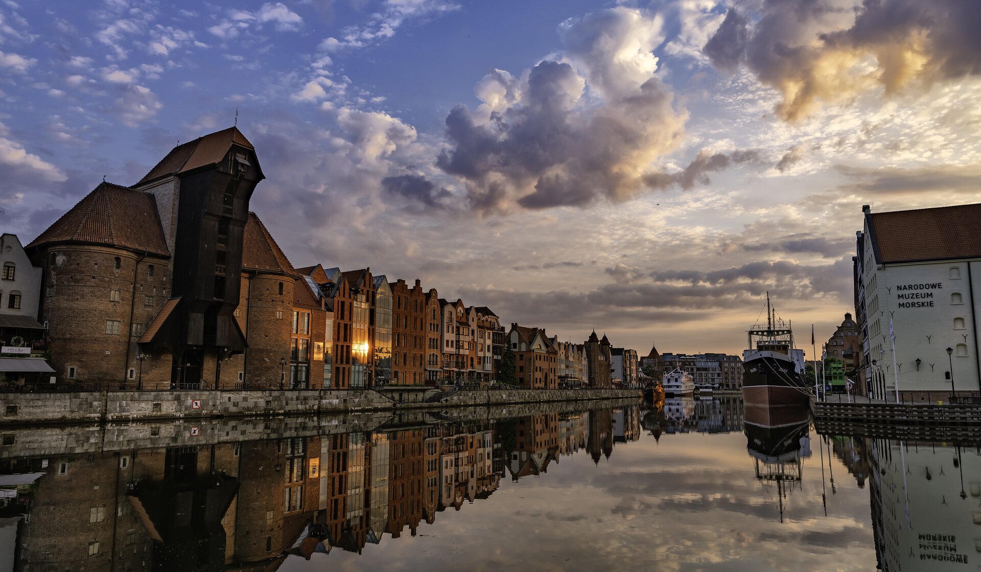 Fotografia przedstawia budynki usytułowane wzdłuż kanału, po jego obu stronach. Z lewej zabytkowy żuraw z drewnianą fasadą. Po prawej stronie jest biały budynek Narodowego Muzeum morskiego, przy brzegu czarno‑biały statek. W oddali po lewej stronie biały statek. Niebo jest zachmurzone. Panuje półmrok.