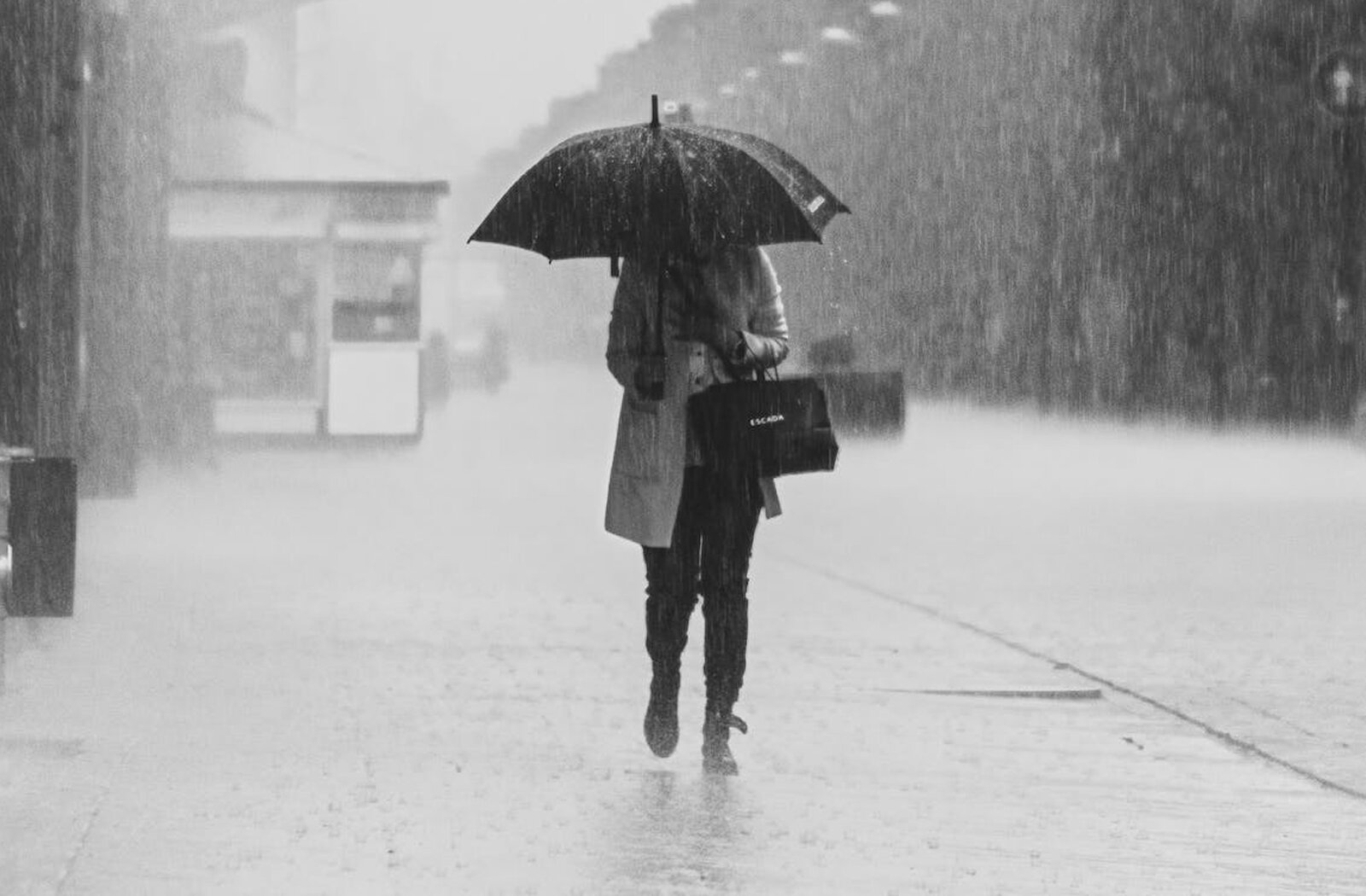 Czarno‑białe zdjęcie przedstawia postać idącą chodnikiem w deszczu. Postać ubrana jest w jasny płaszcz, ciemne spodnie, wysokie ciemne buty, na przegubie lewej ręki ma zawieszoną czarną torbę, a w prawej ręce trzyma duży, czarny parasol, którym zakrywa głowę. Na prawo od postaci widoczna jest ulica, a w tle widoczne są budynki i padający ulewny deszcz.