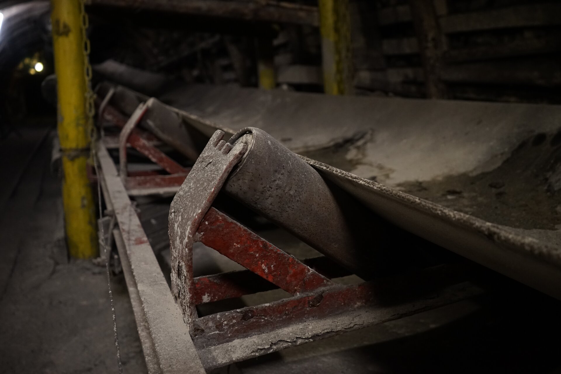 Na zdjęciu widać fragment przenośnika taśmowego czyli urządzenia wykorzystywanego do transportu materiału na dłuższe odległości z podstawowymi jego podzespołami czyli krążnikami, taśmą oraz kozłem górnym. Krążniki są to obrotowe wałki, po których przesuwa się taśma przenośnika.Przenośnik taśmowy jest to podłużne urządzenie mechaniczne na górze którego znajduje się czarna taśma. Podstawą przenośnika taśmowego jest stalowa konstrukcja kratowa czyli trasa. Konstrukcja ta zbudowana jest powtarzalnych członów modułów których podstawą są ceowniki. Ceownik jest to wyrób hutniczy, walcowany lub gięty z metalu. Wyroby te są w postaci prostych odcinków. Przekrój poprzeczny czyli profil ceownika przypomina literę C.Z boku konstrukcji czyli na boku trasy znajdują się metalowe elementy w kształcie ceowników, które biegną pionowo w górę. Końcówki tych elementów są zakrzywione w kierunku taśmy czyli w kierunku środka przenośnika. Są to kozły górne. Kozły górne są rozmieszczone po oby stronach trasy. Na koźle górnym widać krążnik górny. Krążnik jest w kształcie czerwonego walca. Na krążnikach znajduje się długa taśma która biegnie przez całą długość przenośnika.Od boku trasy odchodzą równomiernie stalowe elementy  w kształcie odwróconej litery L. Przedstawiony na zdjęciu przenośnik znajduje się w kopalni. Z prawej strony zdjęcia widać obudowę wyrobiska.