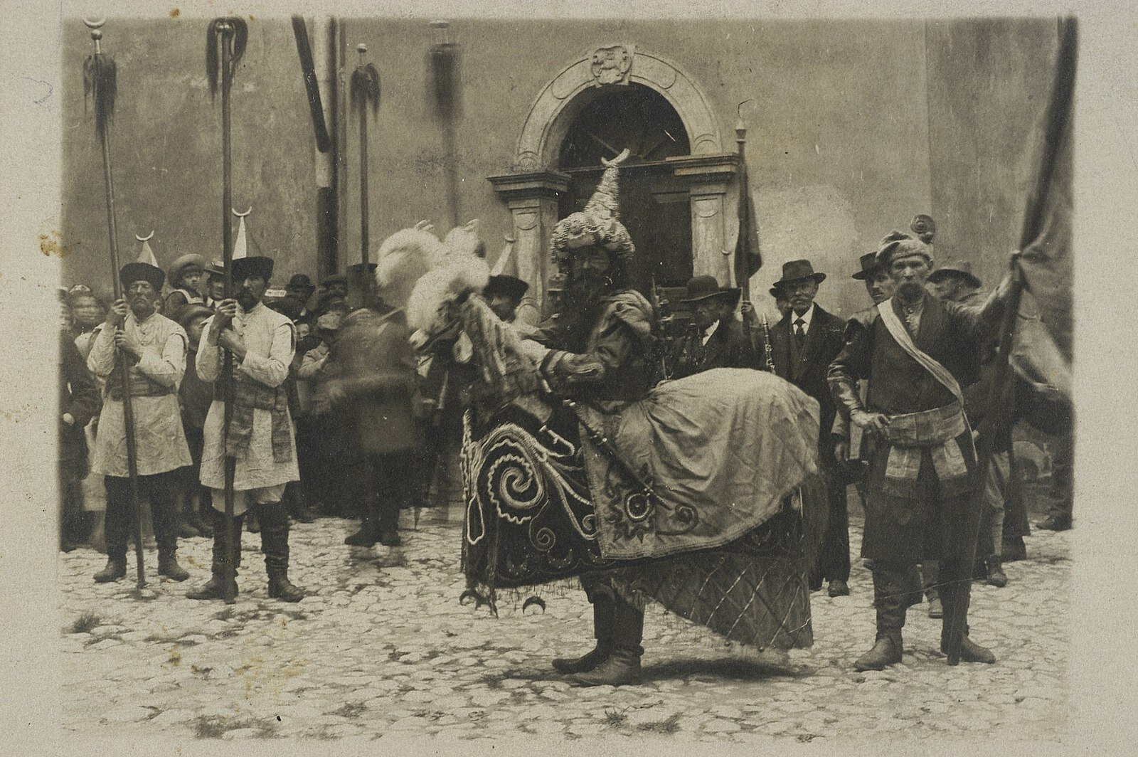 Kliknij, aby powiększyć. Czarno‑białe zdjęcie przedstawia Lajkonika. Ma długą, ciemną brodę. Ubrany jest w bogato zdobiony strój, spiczastą czapkę i wysokie buty. Do pasa postaci przytwierdzony jest sztuczny koń z pióropuszem na głowie, którego grzbiet ozdobiony jest bogato zdobionym, wyszywanym materiałem. W tle widać ludzi przebranych w dawne stroje tatarskie (wysokie buty, spiczaste czapki, długie koszule przewiązane szerokimi szarfami), część z nich trzyma w dłoniach chorągwie. W tle widnieje wejście do budynku.