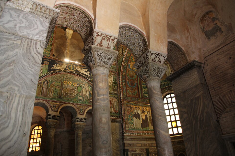 Fotografia przedstawia wnętrze budynku, którego konstrukcja została oparta na kolumnach z głowicami koszowo nasadnikowymi, które mają kształt graniastosłupa prostego o podstawie trapezu prostokątnego. Węższa część głowicy styka się z kolumną, szersza z łukiem, który stanowi część konstrukcji dachowej. Ściany głowicy mają zdobienia ażurowe, w formie okręgów. Wnętrze budynku jest bogato zdobione - pełno w nim obrazów przedstawiających świętych oraz fresków.