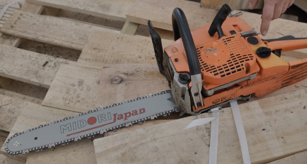 Zdjęcie przedstawia piłę spalinową. Z jednej strony posiada ona uchwyt, a z drugiej prowadnica z łańcuchem z ząbkami.