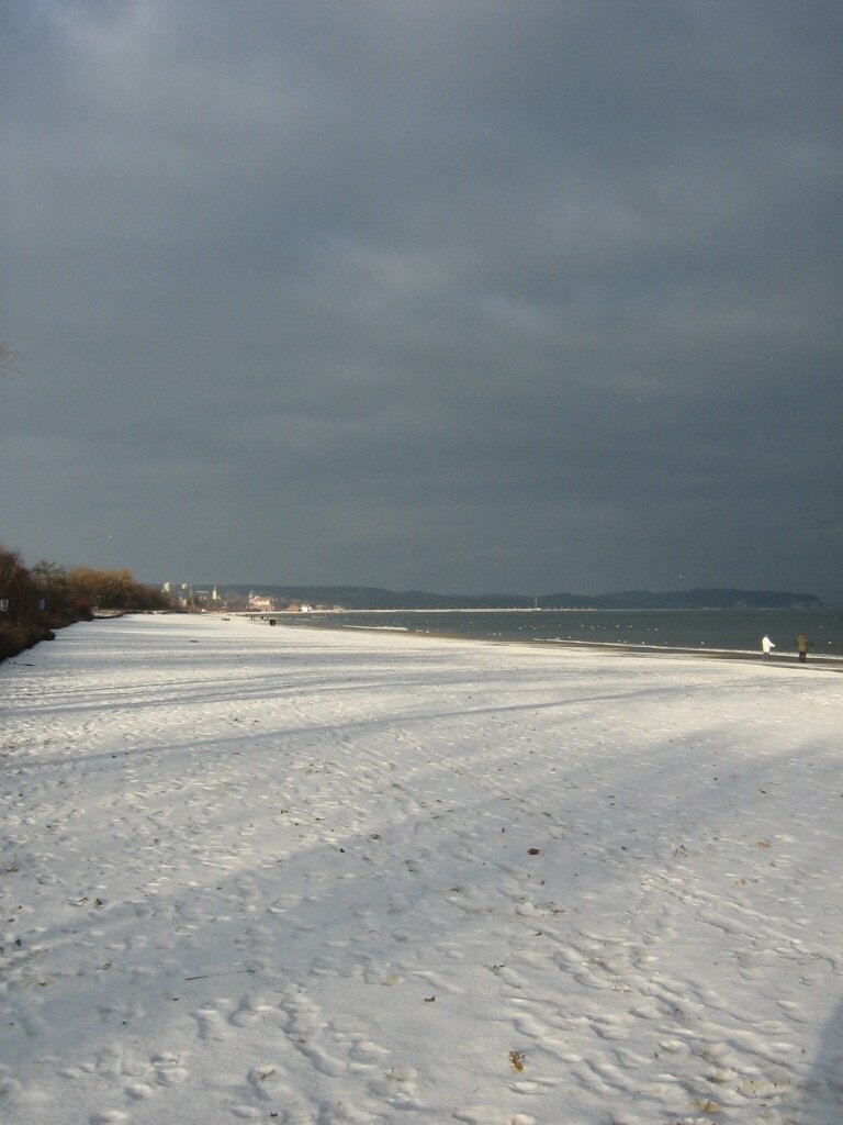 Fotografia przedstawiająca krajobraz zimowy. Na pierwszym planie warstwa śniegu na ziemi. Niebo jest zachmurzone. Po prawej znajduje się zbiornik wodny. 