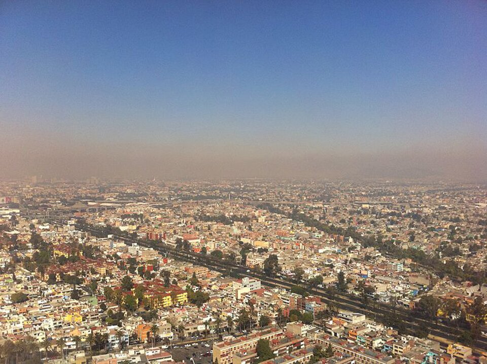 Zdjęcie przedstawia panoramę miasta ukazaną z lotu ptaka. Nad miastem unosi się smog. Powietrze nie jest przejrzyste, jest w kolorze szaro‑różowym.
