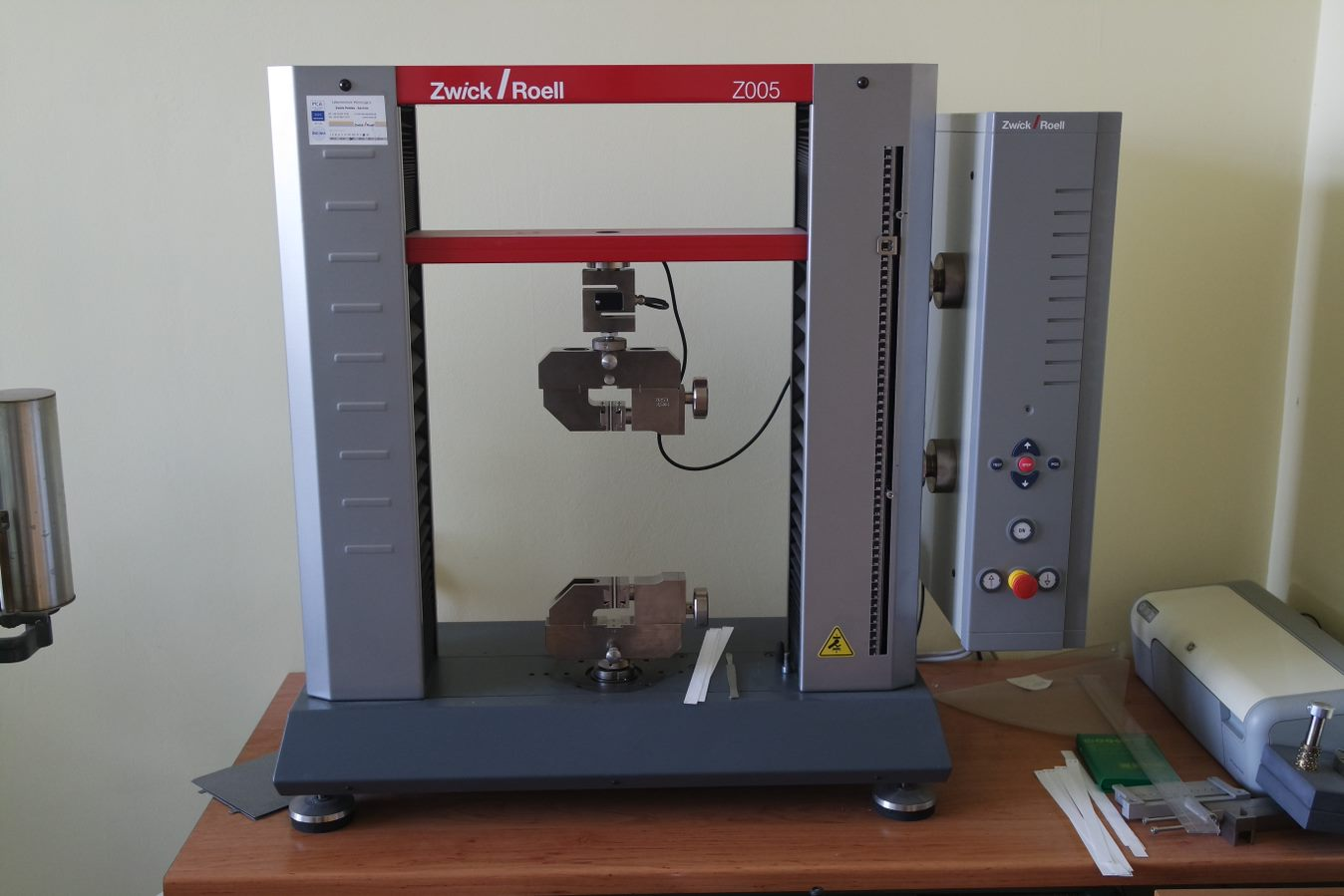 Zdjęcie przedstawia zrywarkę do oznaczania właściwości wytrzymałościowych. Widoczne jest urządzenie składające się z dwóch pionowych elementów, które stanowią oparcie dla środkowego poziomego elementu. Z niego zwisa w dół kolejny element. Pod nim znajduje się drugi taki sam element. Są to szczęki.