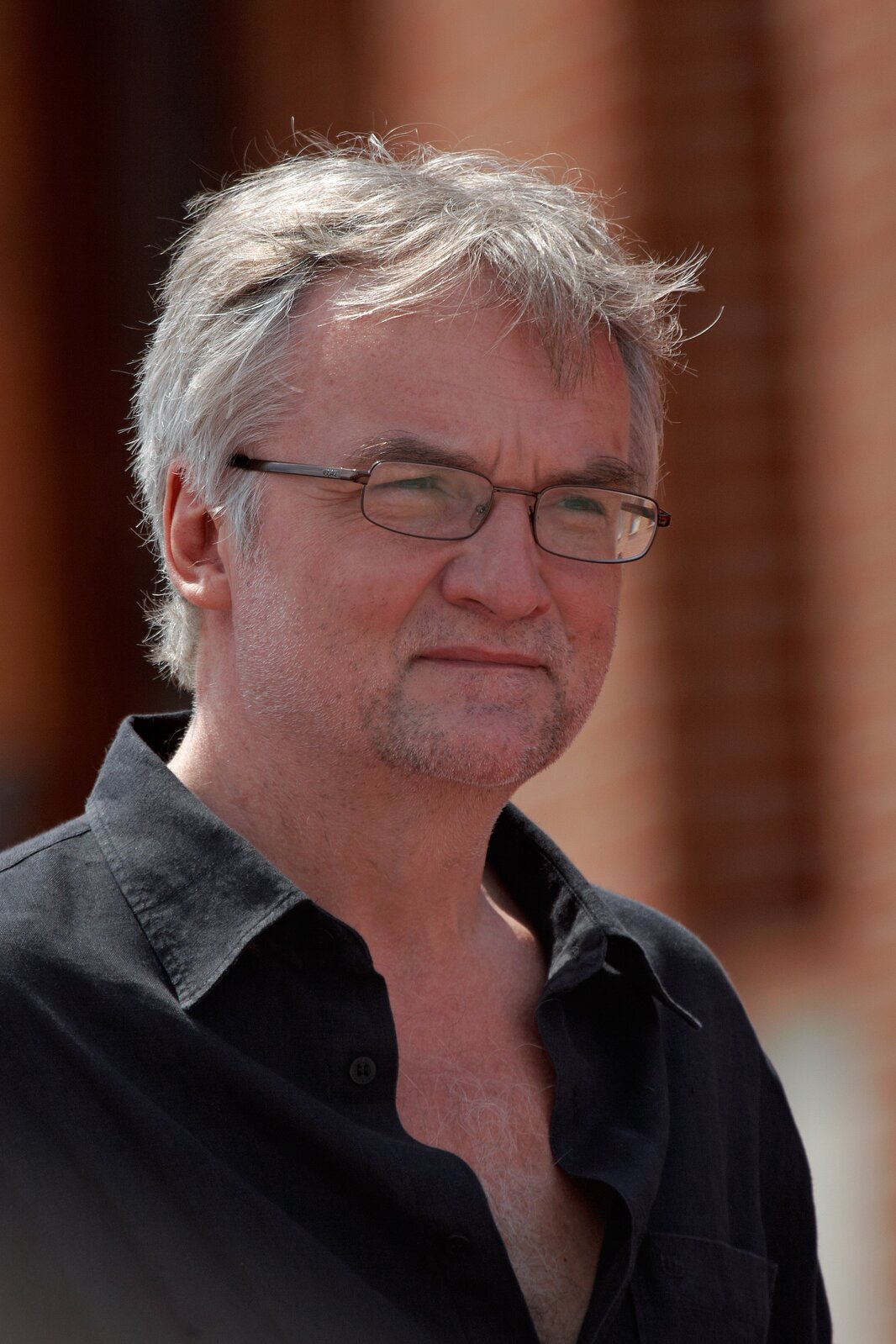 Zdjęcie przedstawia mężczyznę w średnim wieku w okularach i siwych włosach. W ciemnej koszuli rozpiętej pod szyją. Tło fotografii jest nieostre.