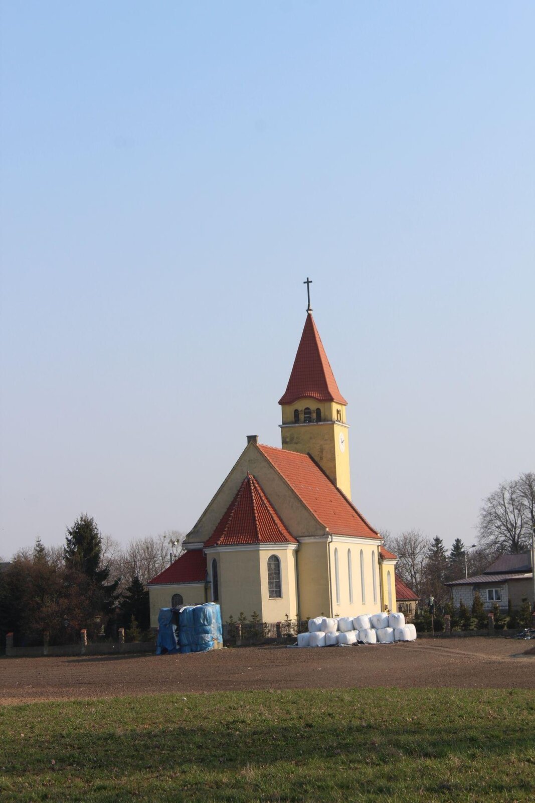 Zdjęcie przedstawia żółty kościół z pomarańczowym dachem. Z przodu kościoła widoczna jest wysoka wieża, na szczycie której znajduje się krzyż. Zdjęcie ukazuje kościół od tyłu, od strony ołtarza. Z obu stron kościoła widoczne są choinki i bezlistne drzewa. Za kościołem jest pole, które częściowo jest pokryte piaskiem, a następnie zieloną trawą. Niebo jest błękitne, bezchmurne. Zdjęcie zostało wykonane w świetle dziennym.
