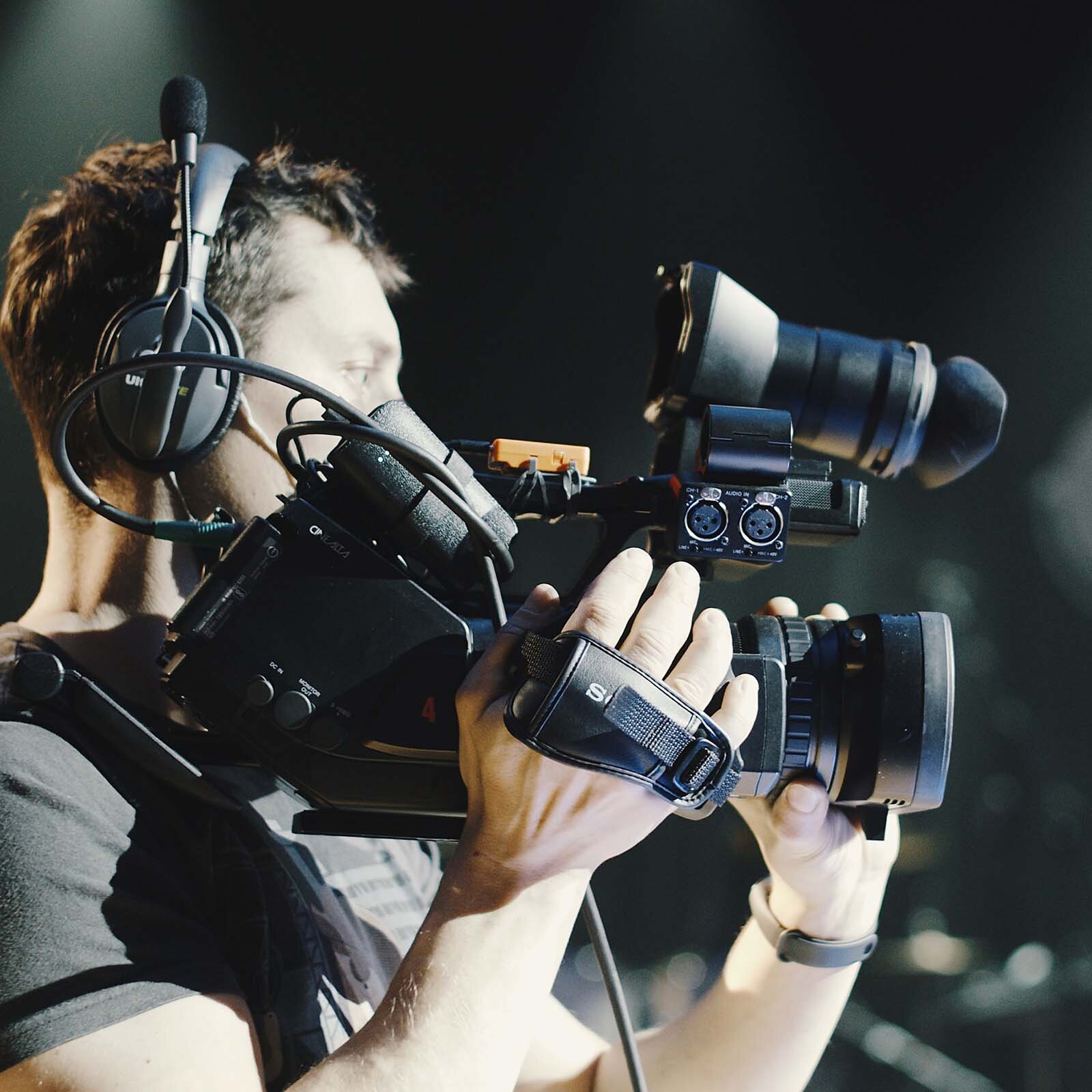 Zdjęcie przedstawia mężczyznę od łokcia w górę, w dużych nausznych słuchawkach, trzymającego profesjonalną filmową kamerę na ramieniu.