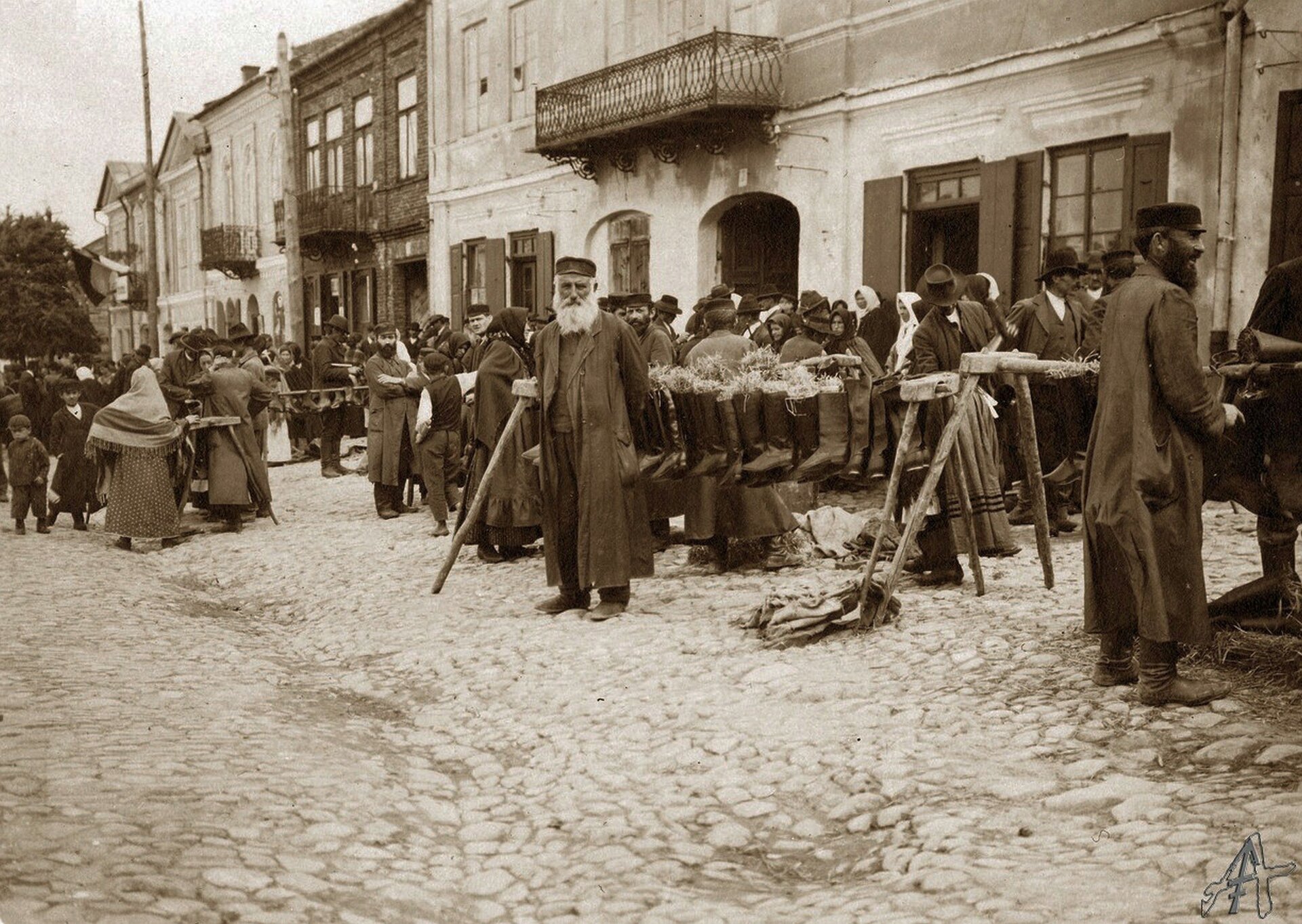 Fotografia przedstawia brukowaną ulicę w niewielkim mieście. Po jej prawej stronie ustawione są liczne kramy. Stoją przy nich kupcy żydowskiego pochodzenia. Są to brodaci mężczyźni w różnym wieku. Ich głowy przykrywają czapki. Są ubrani w długie, ciemne szaty. Wśród stoisk chodzą liczni kupujący. Wśród nich są głównie kobiety. Mają głowy okryte chustami. W tle stoją podniszczone dwukondygnacyjne kamienice.