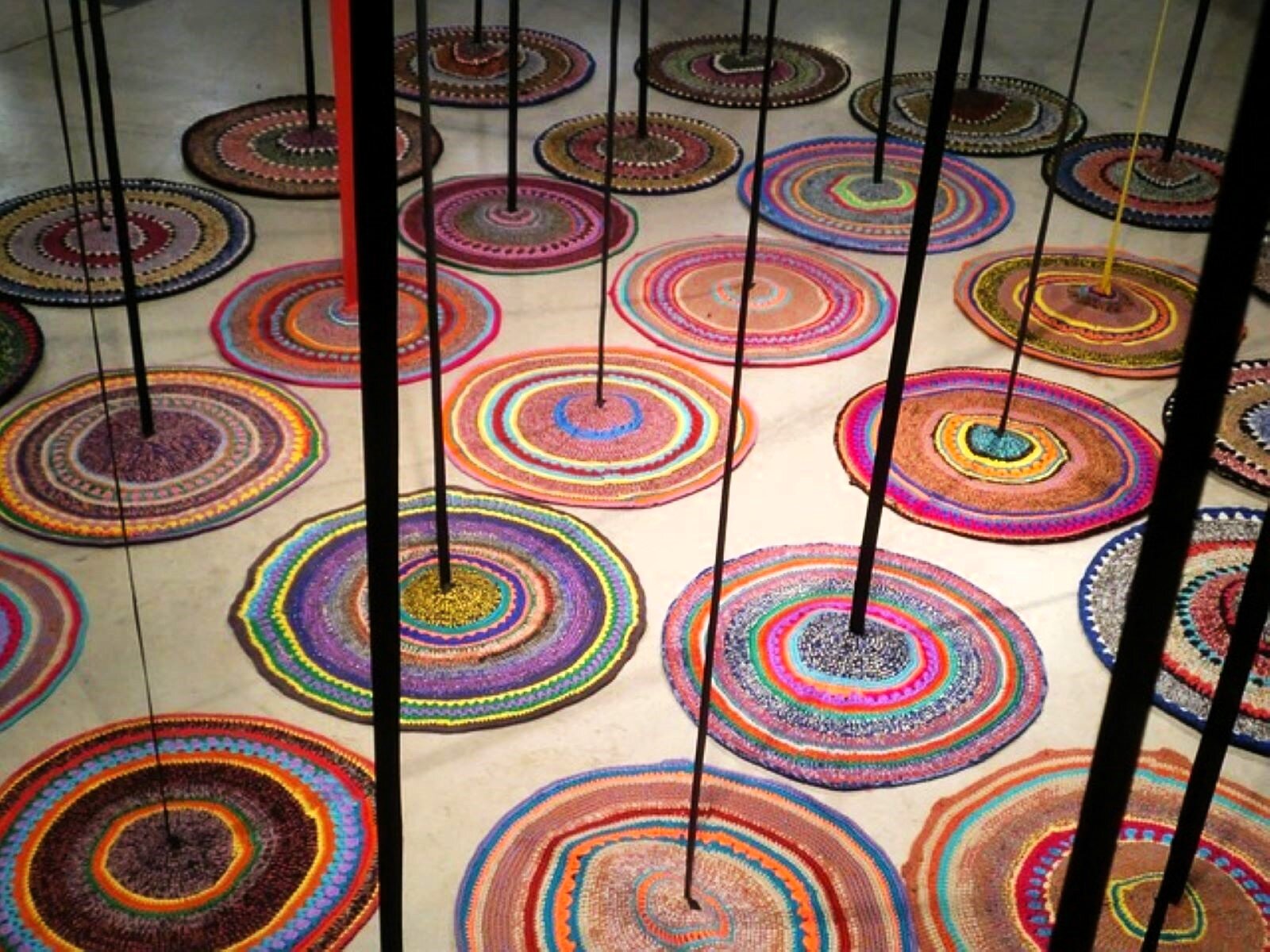 Zdjęcie ukazuje instalację, złożoną z okrągłych dywaników zrobionych na szydełku. Dywaniki leżą obok siebie na podłodze galerii, a do środka każdego z nich opuszczona jest pionowo z góry wąska taśma, która przypomina pień drzewa. 
