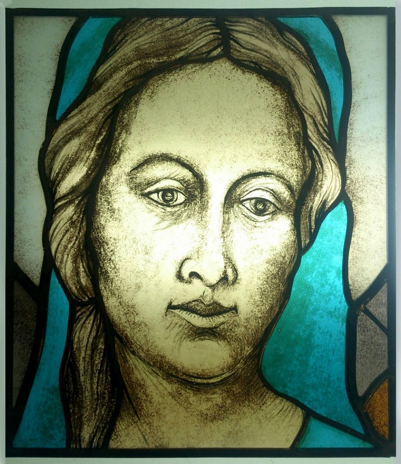 Zdjęcie przedstawia fragment witraża Witolda Pochylskiego "Święta Rodzina". Ukazuje głowę Matki Boskiej. Charakteryzacje się rysunkiem twarzy , na której został zaakcentowany światłocień. Głowę Marii pokrywa błękitny płaszcz.