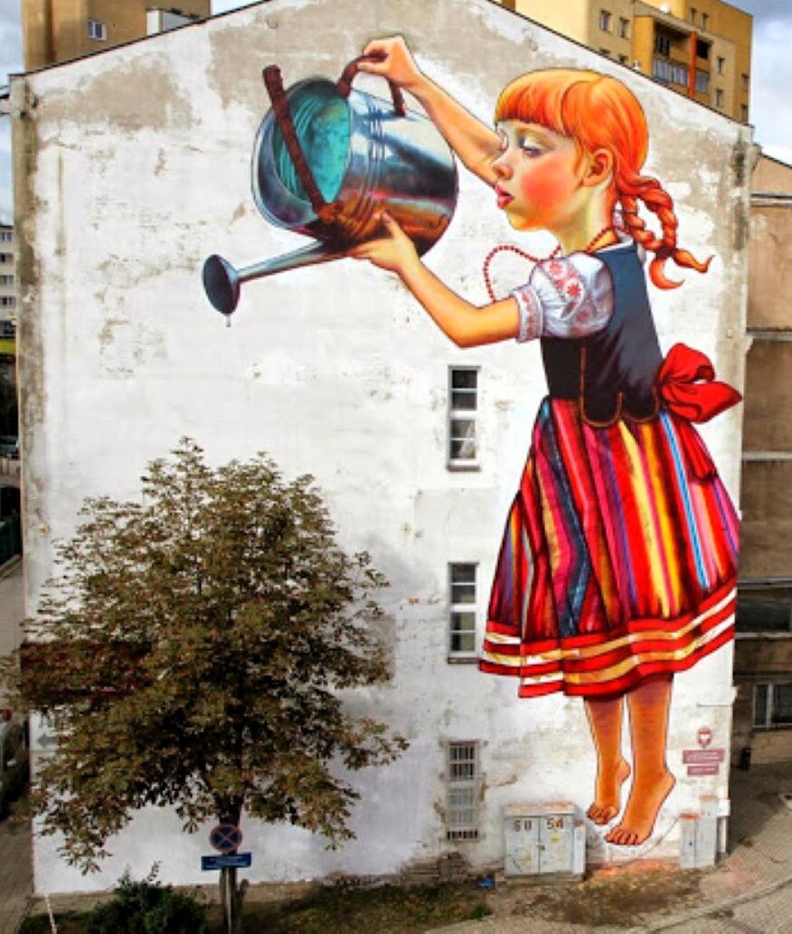 Ilustracja przedstawia mural na bocznej ścianie wielopiętrowego budynku. Na białym tle jest postać dziewczynki z warkoczami, w kolorowym stroju ludowym, która przechylając konewkę podlewa z góry drzewo. Drzewo nie jest namalowane - rośnie przy ścianie budynku.