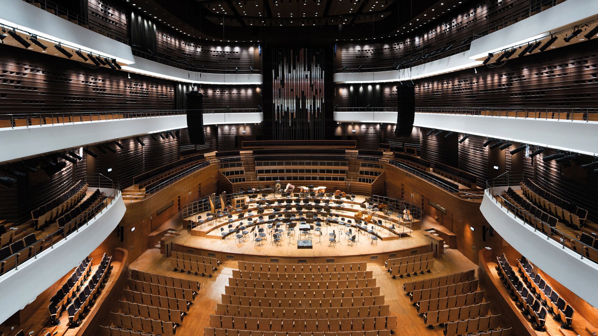 Fotografia przedstawiająca salę koncertową w budynku Narodowego Forum Muzyki we Wrocławiu. W głębi usytuowana jest scena dla miejscami dla muzyków i dyrygenta. Za nią miejsca dla chóru. Na poziomie sceny, przed nią jest pierwszy poziom miejsc dla widowni. Wokół niego umiejscowiono balkony na trzech poziomach.