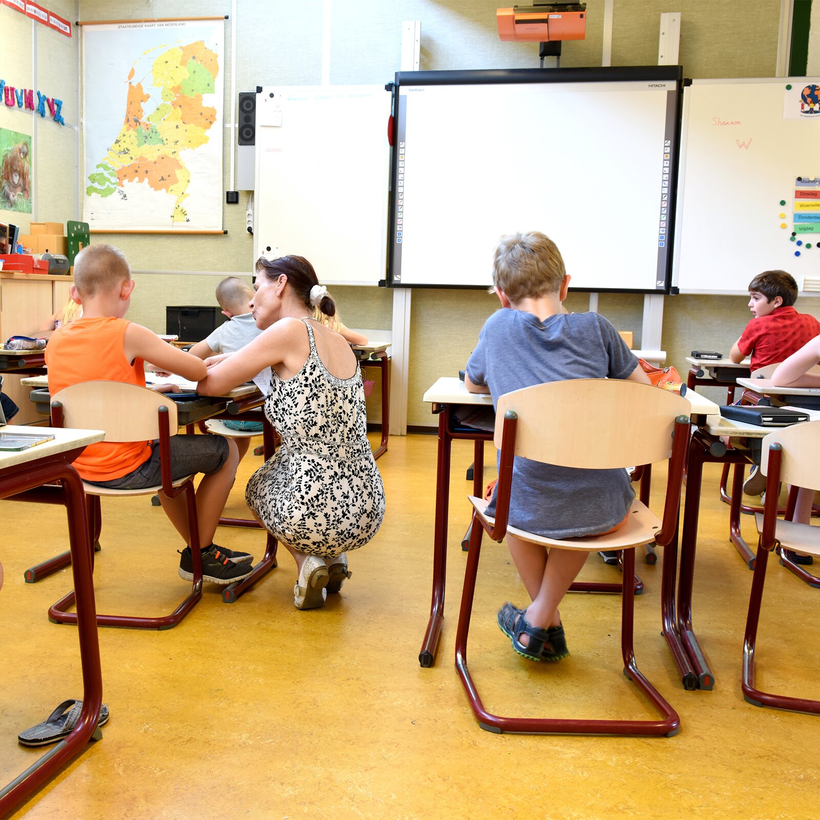 Ilustracja przedstawia dzieci siedzące w klasie w pojedynczych ławkach na krzesłach. Przy jednym z nich kuca nauczycielka. Z przodu widoczna jest tablica i pomoce szkolne. Na ścianie wisi mapa, obrazki ze zwierzętami oraz litery alfabetu. Nad tablicą wisi projektor. Przy ścianach stoją szafki. Uczniowie na biurkach mają tablety.