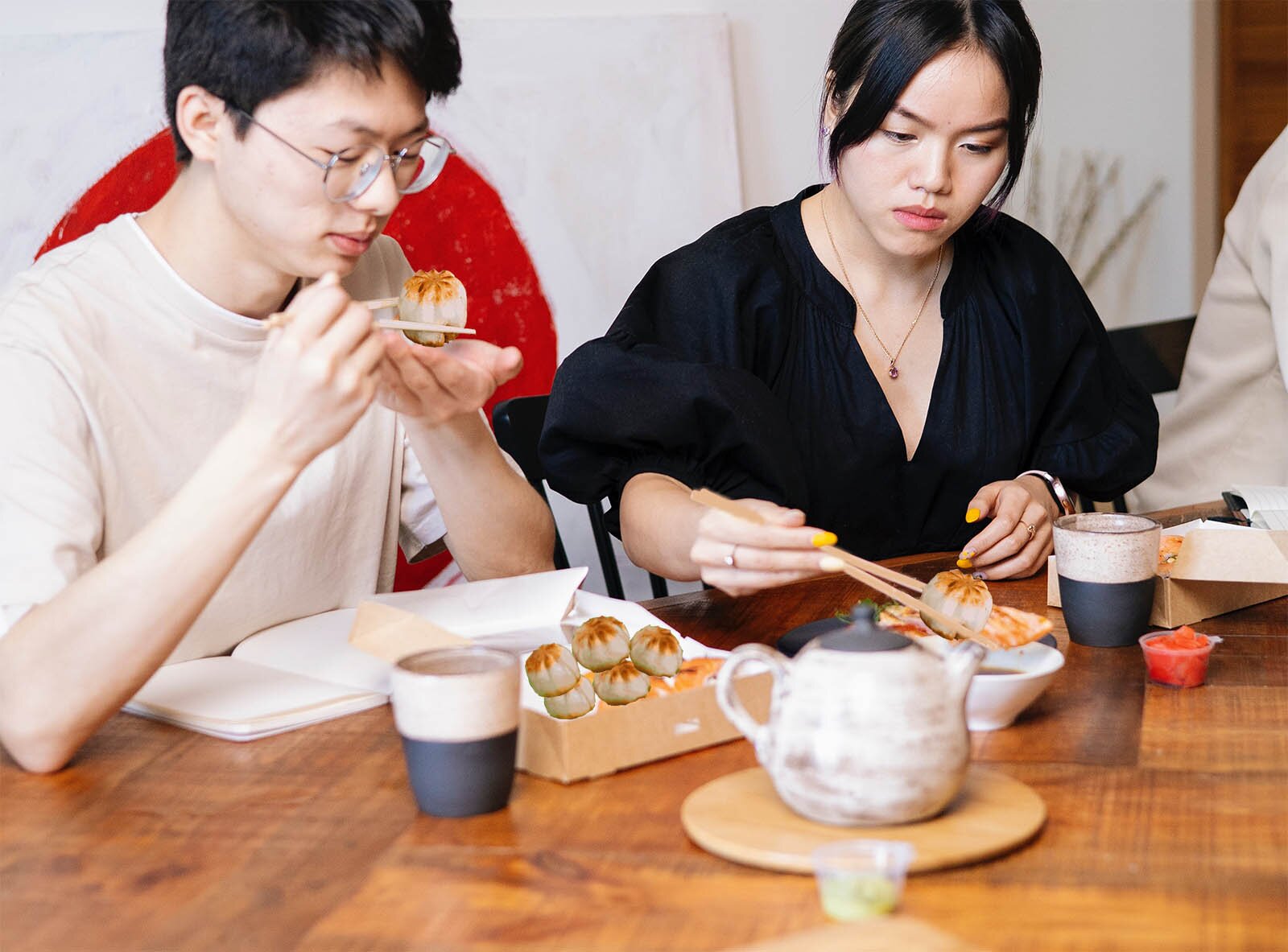 Zdjęcie przedstawia dwoje młodych ludzi siedzących przy stole jedzących pałeczkami okrągłe pierożki. 