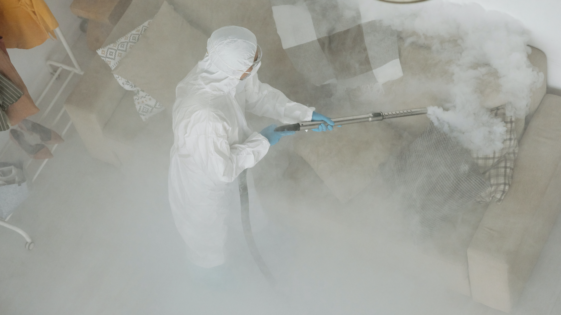 Zdjęcie przedstawia pracownika, który wykonuje dezynfekcję w pomieszczeniu. Ubrany jest w specjalistyczny kombinezom, który zakrywa całe ciało. W ręku trzyma urządzenie do wykonywania dezynfekcji.