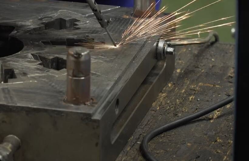 Ilustracja przedstawia powierzchnie formy odlewniczej, w trakcie nakładania powłoki wolframowo molibdenowej. Widoczna jest końcówka urządzenia do nakładania powłoki, stykająca się z powierzchnią formy. W miejscu styku, powstają iskry strzelające we wszystkich kierunkach.