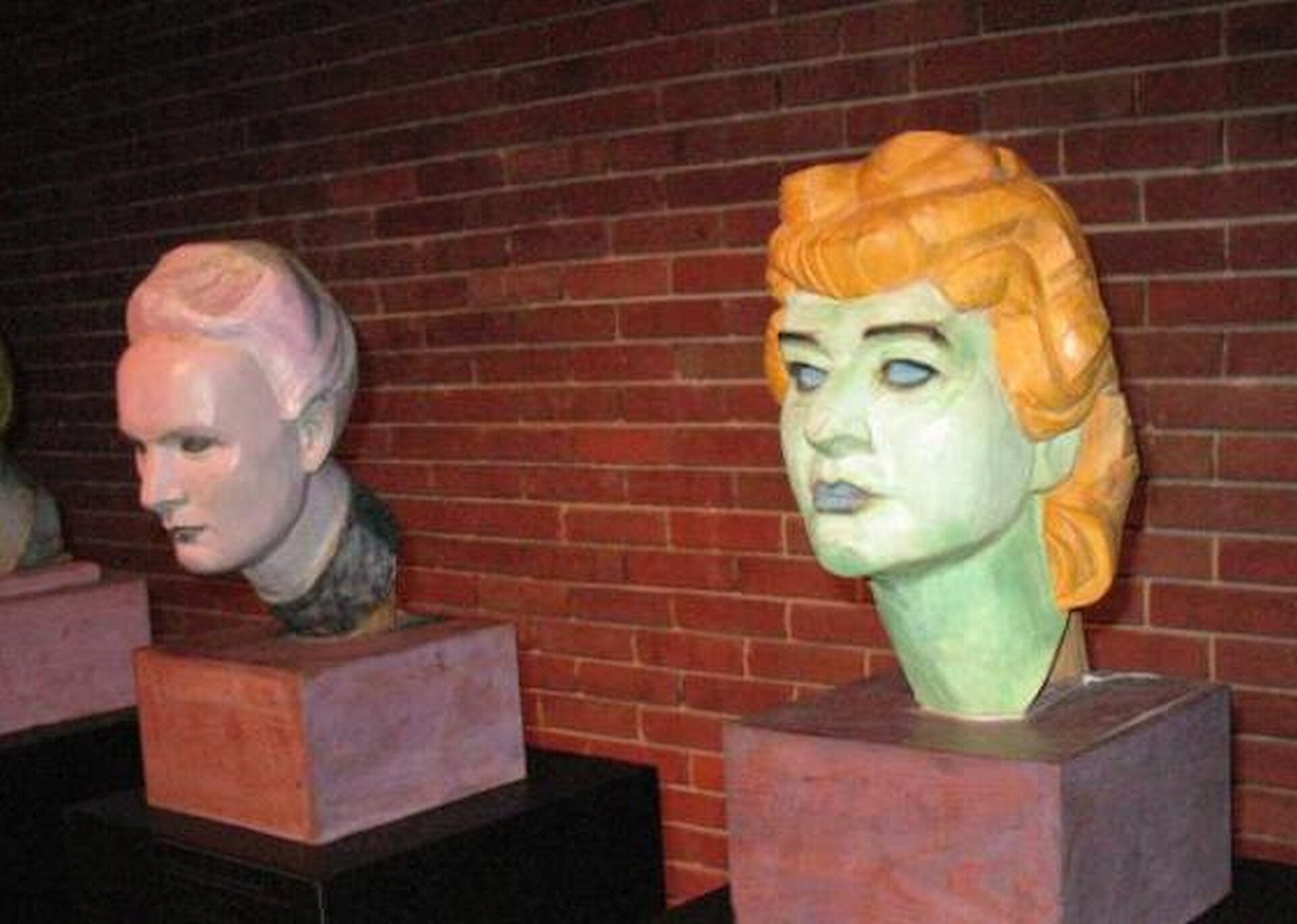 Ilustracja przedstawia dwie rzeźby Helena Modrzejewska, z drugiego cyklu Głowy wawelskie, Xawery Dunikowski, Muzeum Narodowe w Warszawie. Na ilustracji widać dwie rzeźby głowy. Na pierwszym planie widać głowę w kolorze lekko zielonym z pomarańczowymi włosami, ułożonymi w fale. Oczy są szeroko otwarte, wydatnie zarysowany nos i usta. Druga rzeźba to głowa starszej kobiety z siwymi włosami upiętymi w górę. rzeźby ustawione są na tel muru z czerwonej cegły.