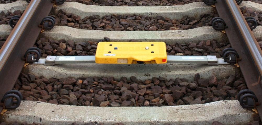 Zdjęcie przedstawia żółtą baliasę zamontowaną pośrodku torowiska. Jest to małe, płaskie, prostokątne urządzenie, zamontowane na metalowych wspornikach.