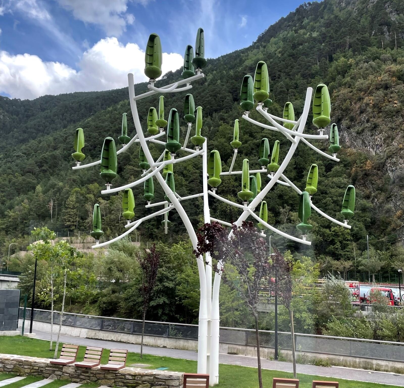 Zdjęcie przedstawia instalację w naturalnej przestrzeni. Jest to drzewo wykonane z pomalowanego metalu i sztucznego tworzywa. Pień i gałęzie są neutralne w kolorze, a na gałęziach zostały umieszczone stojące jakby pąki ze sztucznego tworzywa, w kolorze trawy i liści. Wydaje się to nierealnym zestawieniem w stosunku do otoczenia żywych drzew i naturalnej górskiej roślinności.  