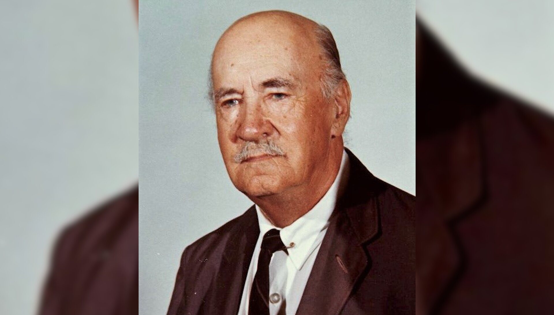 Fotografia przedstawiająca portret starszego mężczyzny z wąsem, ubranego w koszulę, krawat i marynarkę.