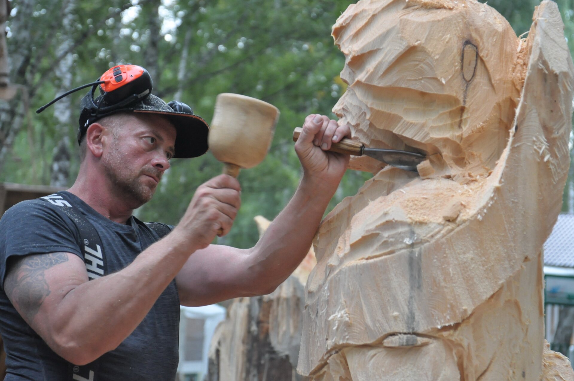 Ilustracja interaktywna przedstawia mężczyznę rzeźbiącego w drewnie w czasie festiwalu Ax Day, który odbywa się na świeżym powietrzu. Jest on w średnim wieku, jego krótkie włosy są brązowo‑siwe. Na jego twarzy widoczny jest krótki zarost. Ma na sobie koszulkę odsłaniającą tatuaż na prawej ręce, w której trzyma pobijak, w drugiej dłoni trzyma dłuto. Mężczyzna bije pobijakiem w dłuto, które trzyma przy fragmencie drewna. Na głowie mężczyzna ma czapkę oraz słuchawki wygłuszające dźwięki, nie są one jednak nasunięte na jego uszy. Tło stanowi las oraz inne drewniane rzeźby. 