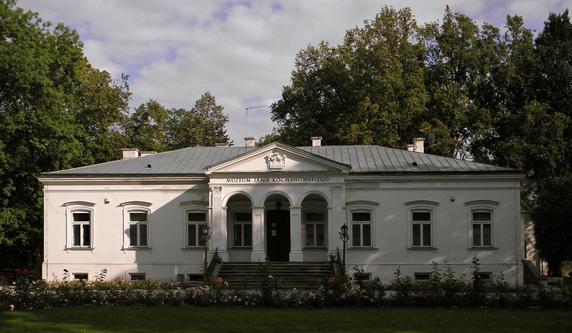 Fotografia barwna przedstawiająca biały dworek otoczony drzewami. Jest to niski budynek, który przy wejściu ma cztery kolumny zwieńczone trójkątną płaskorzeźbą.