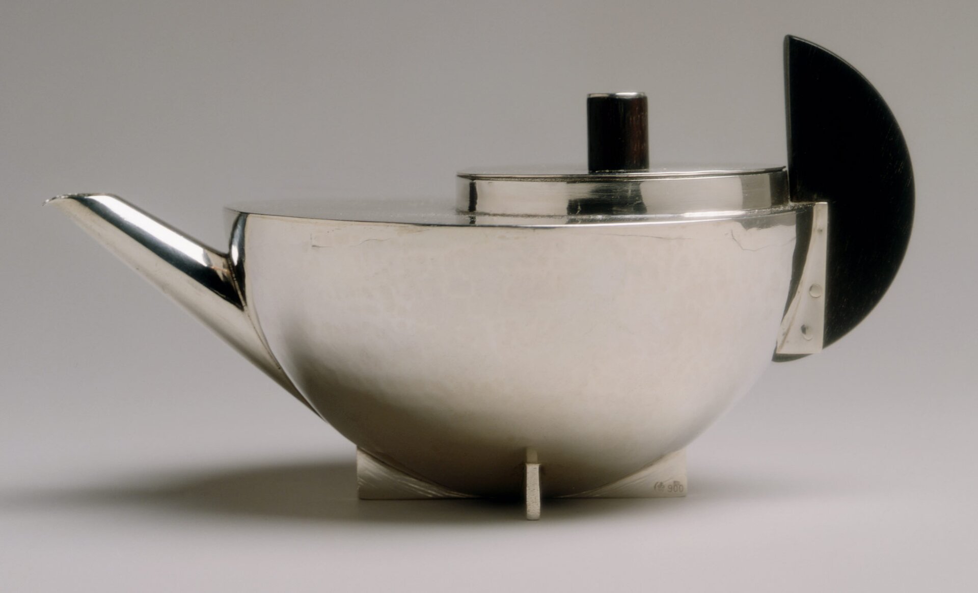 Fotografia przedstawia srebrny imbryk do herbaty. Naczynie wykonane jest ze stali nierdzewnej o kształcie półkuli z prostym wylewem i okrągłą pokrywką, wystającą na jednej połowie płaskiej części czaszy imbryka. Uchwyt jest czarnym półksiężycem.