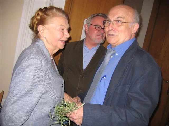Kliknij, aby powiększyć Na zdjęciu znajdują się trzy osoby. Z lewej strony starsza kobieta z włosami upiętymi w kok w jasnej marynarce, uśmiecha się – jest to Julia Hartwig. Po prawej stronie kobiety stoi starszy mężczyzna w okularach, w ciemnej kurtce i koszuli z krawatem – to Ryszard Kapuściński. Między nimi w oddali stoi mężczyzna w ciemnej marynarce w okularach i z zarostem – to Anders Bodegård. W tle znajdują się drzwi.