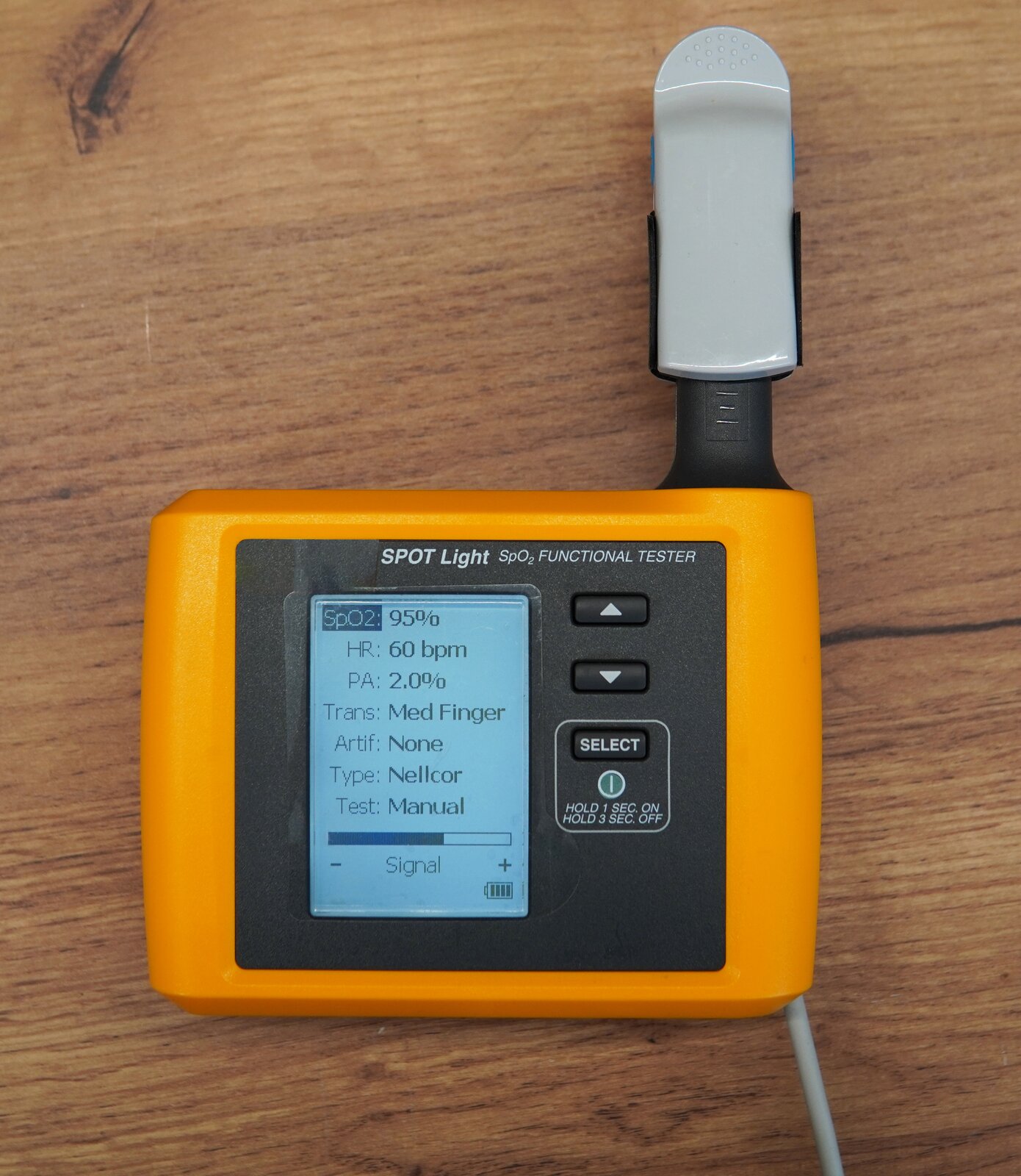 Zdjęcie przedstawia symulator nasycenia krwi tlenem SpO2. Na górnej ścianie obudowy znajduje się panel sterujący z przyciskami oraz wyświetlacz z informacjami o mierzonych wartościach. Do urządzenia podłączony jest pulsoksymetr. 