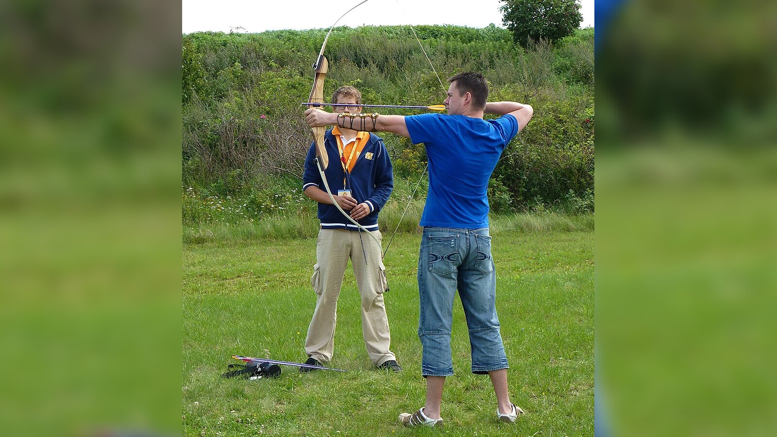 Zdjęcie przedstawia dwóch mężczyzn w młodym wieku. Jeden z nich trzyma w dłoniach napięty łuk gotowy do strzału. 