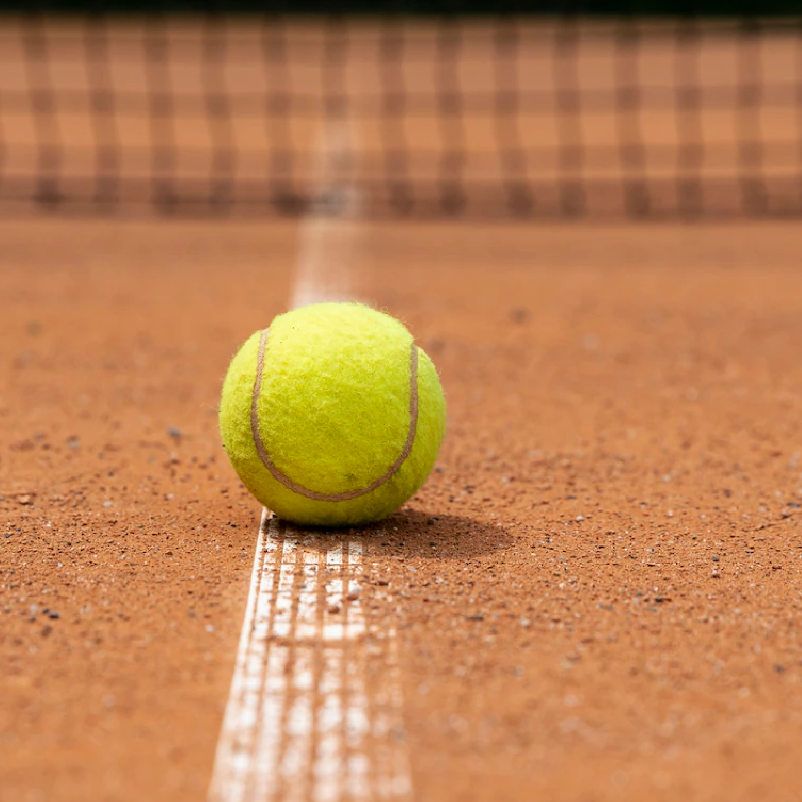 Na zdjęciu żółta piłeczka tenisowa leżąca na linii namalowanej na korcie tenisowym. 