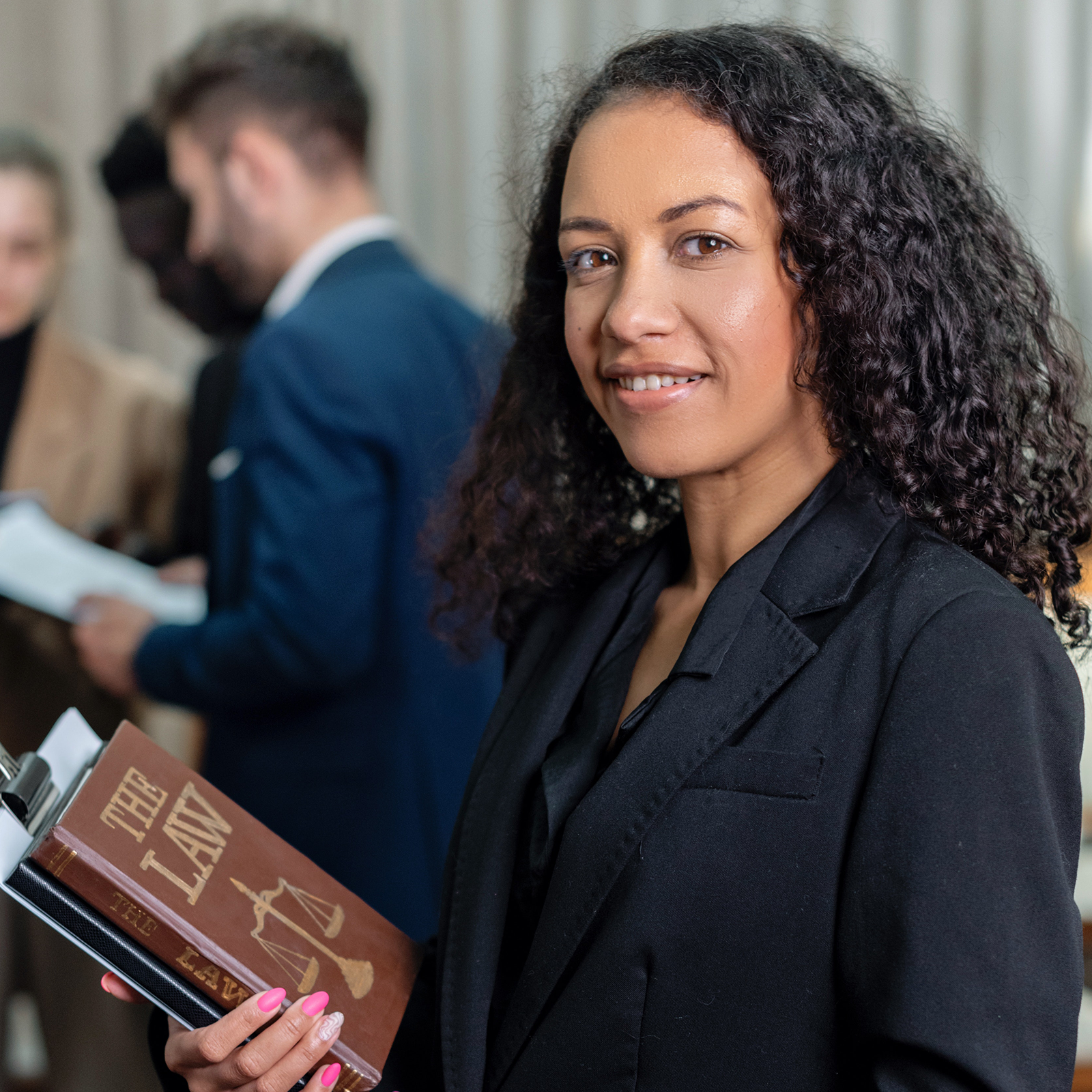 Zdjęcie przedstawia młodą, lekko uśmiechniętą kobietę, ubraną w czarną koszulę i czarną marynarkę. W prawej ręce przy klatce piersiowej kobieta trzyma książki, pierwsza z nich jest zatytułowana "THE LAW".