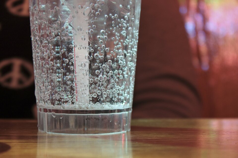 Zdjęcie przestawia stojącą na stole szklankę wypełnioną wodą gazowaną. Woda jest pełna pęcherzyków gazu. Wewnątrz szklanki znajduje się słomka.