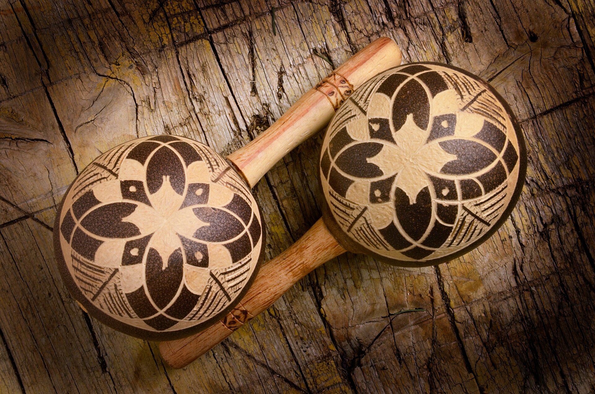 Ilustracja przedstawia dwa drewniane marakasy o kulistym kształcie z podłużnymi uchwytami. Leżą na drewnianej półce i posiadają ozdobny element/wyżłobienia na uchwytach jak i na kulach.