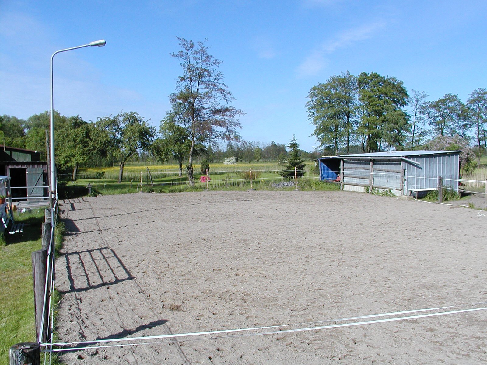 Zdjęcie przedstawia piaszczysty padok. Jest on ogrodzony elastycznymi taśmami. Po lewej stronie widać latarnię, po prawej blaszany barak stojący za ogrodzeniem. W oddali rosną drzewa.