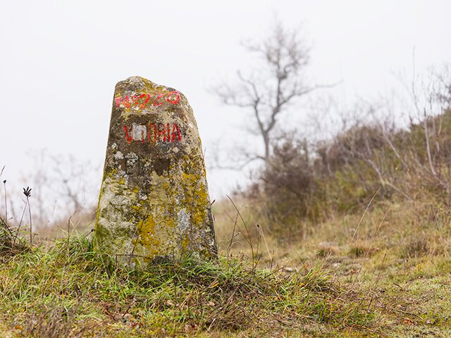 Zdjęcie przedstawia niewysoki kamień stanowiący słupek. Stoi w otoczeniu traw. Pokryty jest żółtym i szarym nalotem. Na kamiennym słupku jest czerwony napis: Victoria.