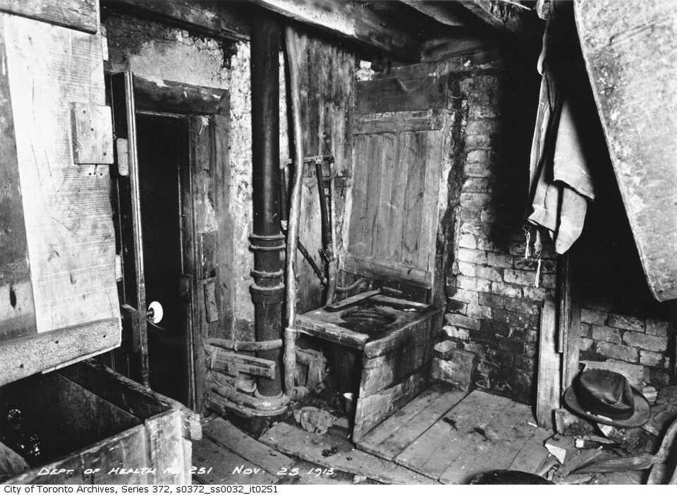 Zdjęcie przedstawia wnętrze mieszkalne w Toronto. Znajdują się w nim nieosłonięte grube rury oraz otwory zabite deskami. Ściany zbudowane są z cegły. Posadzka wyłożona jest deskami.