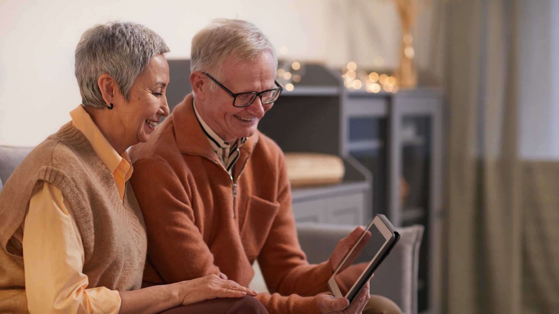 Zdjęcie przedstawia starszą kobietę i starszego mężczyznę. Siedzą na kanapie, są wpatrzeni w ekran tabletu i uśmiechają się.