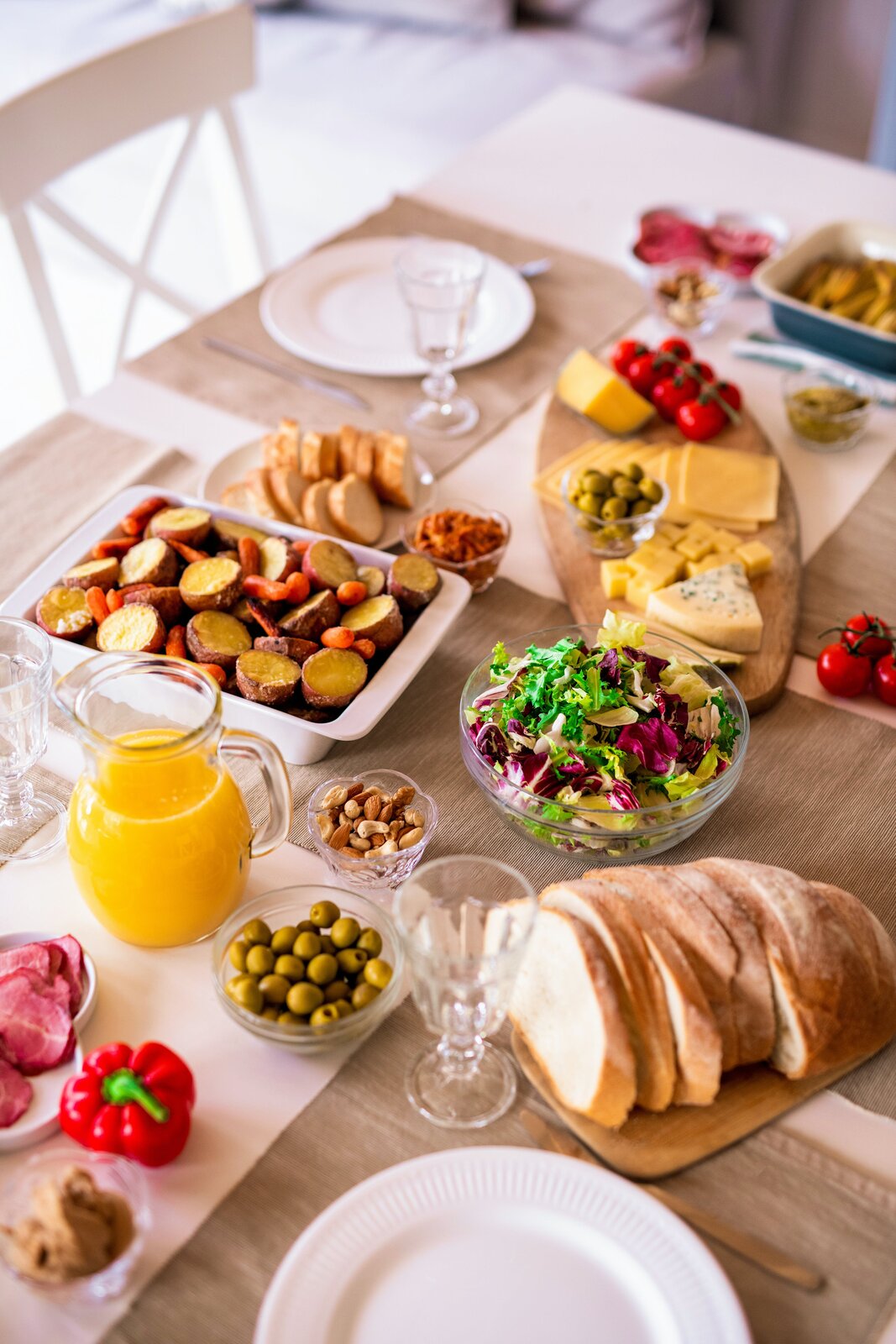 Ilustracja przedstawia zastawiony stół. Stoją na nim między innymi chleb, sery, szynka, owoce, warzywa, sok pomarańczowy, oliwki, orzechy.