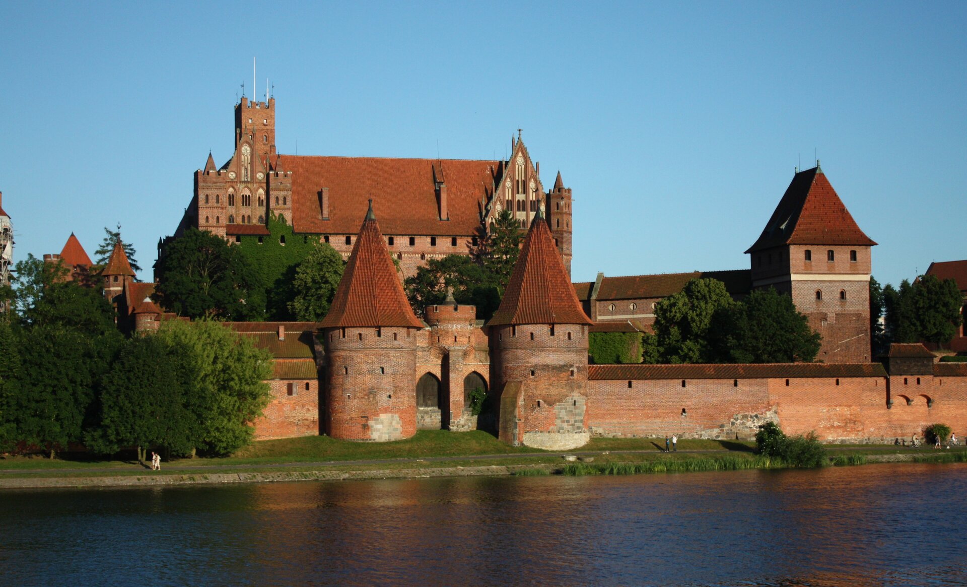 Fotografia barwna przedstawiająca Zamek w Malborku od strony Nogatu - rzeki stanowiącej wschodnie ramię ujściowe Wisły. Color photography depicting the Malbork Castle from the side of the Nogat River - the river constituting the eastern Vistula estuary.